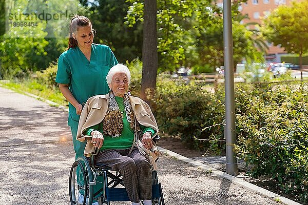 Eine ältere Frau mit der Krankenschwester auf einem Spaziergang im Garten eines Pflegeheims in einem Rollstuhl neben der Natur