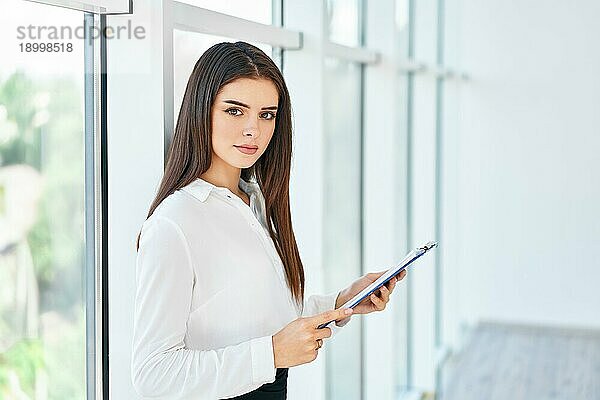 Selbstbewußt lächelnde Geschäftsfrau mit Klemmbrett in einem hellen  modernen Büro mit Kopierraum. Geschäftsleute