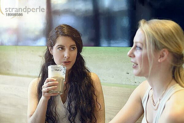 Attraktive  fröhliche  lächelnde junge Frau  die sich mit ihrer Freundin unterhält  während sie an einem Tisch sitzen und gemeinsam einen Drink in einem Restaurant genießen