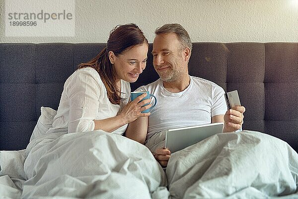 Glückliches Paar mittleren Alters  das morgens im Bett sitzt  Kaffee trinkt und mit einem digitalen Tablet online einkauft