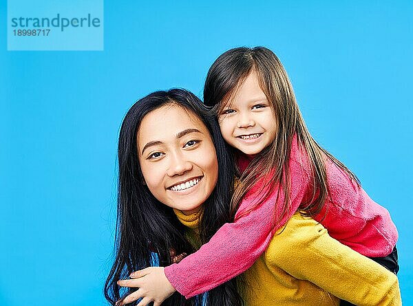 Glückliche Mutter und kleine Tochter Huckepack zusammen vorblauem Hintergrund. Familie Konzept