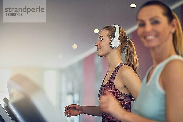 Attraktive junge Frau mit drahtlosen Kopfhörern auf dem Laufband im Fitnessstudio stehend  Seitenansicht und lächelnd mit Kopierraum