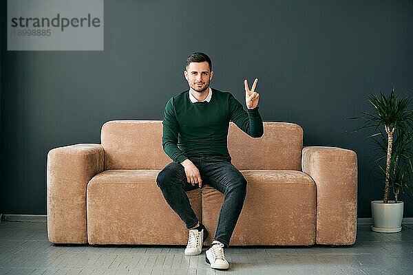 Trendy Mann zeigt Hand Geste V Zeichen für den Sieg oder Frieden suchen ro Kamera beim Sitzen auf moderne Wohnung
