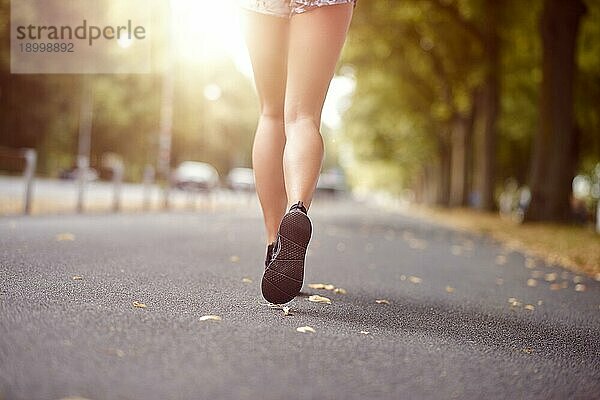 Junge Frau  die eine herbstliche Straße hinuntergeht oder joggt  im Gegenlicht der Abendsonne  in einem niedrigen Blickwinkel auf ihre wohlgeformten Beine in kurzen Hosen und Sandalen von hinten  mitten im Schritt