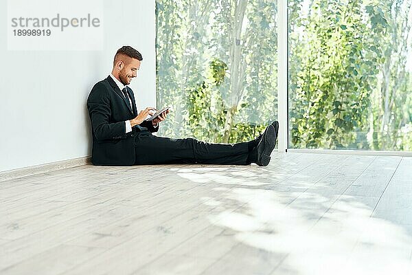 Junge lächelnde Geschäftsmann sitzt auf dem Boden in seinem Büro und Blick auf Smartphone. Pause Zeit Konzept  Business Technologie