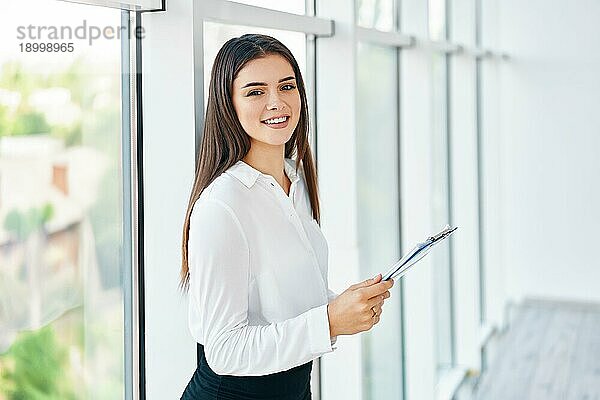 Selbstbewußt lächelnde Geschäftsfrau mit Klemmbrett in einem hellen  modernen Büro mit Kopierraum. Geschäftsleute