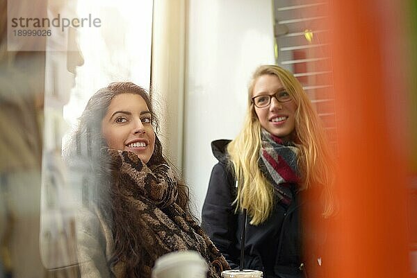 Blonde und brünette Freundinnen in Mänteln unterhalten sich im öffentlichen Raum in der Nähe der orangefarbenen Stange neben dem Fenster