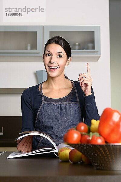 Glückliche Frau mittleren Alters  die eine Schürze trägt und an der Küchentheke mit einem Rezeptbuch steht  mit Betonung auf dem positiven Handzeichen