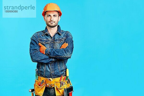 Porträt eines selbstbewussten  gut aussehenden männlichen Bauarbeiters mit verschränkten Armen und Kopierraum  der auf einem blaün Hintergrund steht und zur Kamera schaut