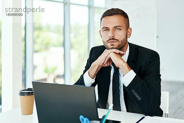 Selbstbewusster junger Geschäftsmann in elegantem Anzug posiert in einem hellen  modernen Büro. sitzt an seinem Schreibtisch. Business Erfolg Konzept
