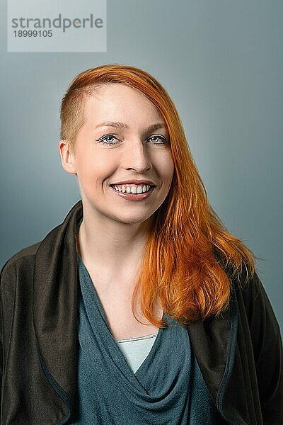 Porträt einer lächelnden  zuversichtlichen rothaarigen Frau mit einer seitlich geschnittenen Frisur  die in die Kamera schaut  auf grauem Studiohintergrund