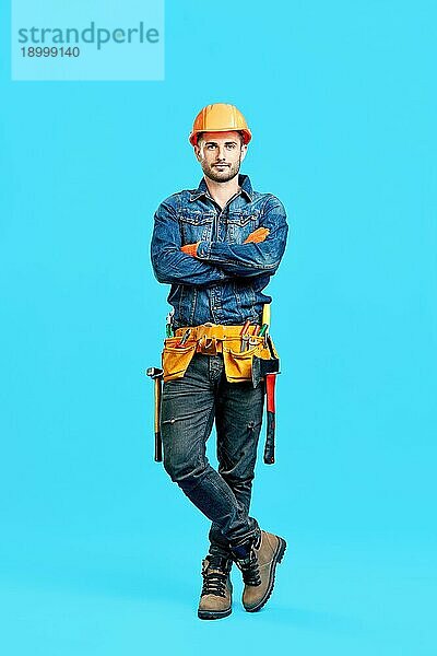 Porträt eines selbstbewussten  gut aussehenden männlichen Bauarbeiters mit verschränkten Armen  der vor einem blaün Hintergrund steht und in die Kamera schaut