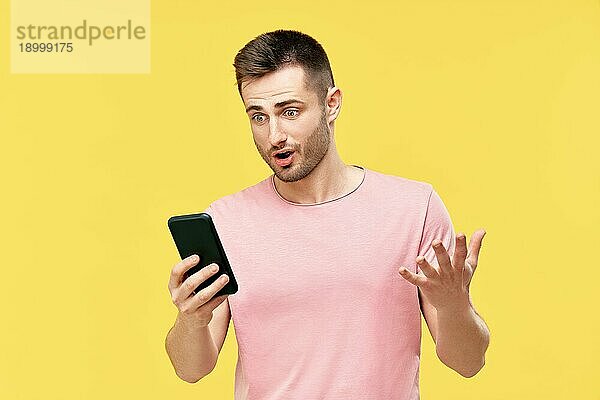 Schockiert lustig Millennial Mann mit Handy hält es in der Hand isoliert über gelben Hintergrund