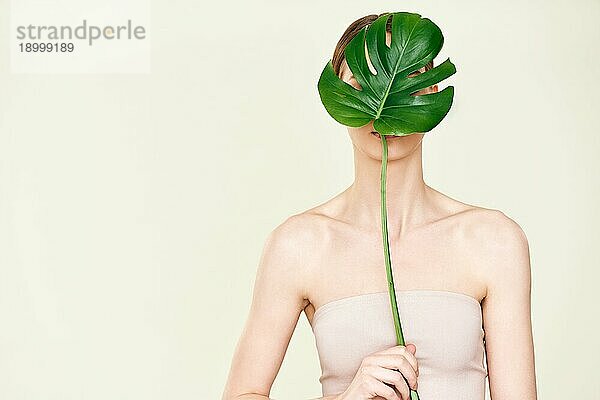 Schöne junge Frau bedeckt und versteckt Gesicht mit monstera tropischen Blatt mit Kopie Raum. Naturkosmetik  Hautpflege  Spa  gesichtslose Person Konzept