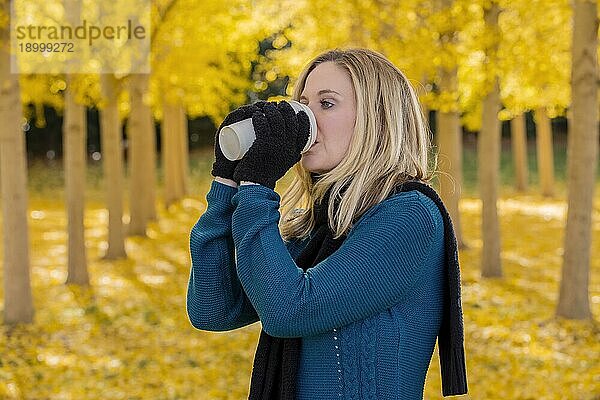 Ein schönes blondes Modell trinkt ein heißes Getränk  während es im Freien in einem Feld mit gelben Blättern posiert