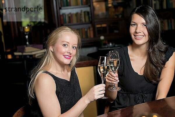 Zwei schöne elegante Frauen Freunde in stilvollen einfachen schwarzen Cocktailkleider Toast einander mit Gläsern von gekühlten Champagner  wie sie auf eine Nacht zusammen feiern