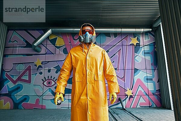 Graffitimaler mit Atemschutzmaske steht in der Nähe der Wand mit seinen Bildern und schaut in die Kamera. Street Art Konzept