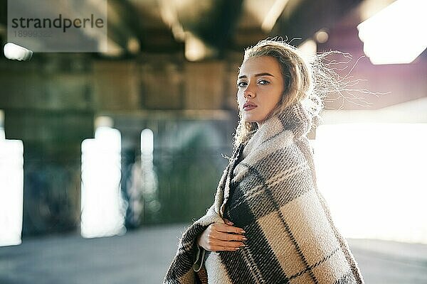Porträt einer hübschen  selbstbewussten Frau  die in eine Wolldecke eingewickelt ist und in die Kamera schaut  posiert auf einem städtischen Straßenhintergrund mit Kopierraum. Ausdruck  weibliche Schönheit  entspannen  Komfort Konzept