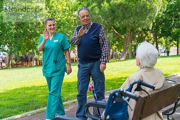 Ein älterer Mann mit der Krankenschwester bei einem Spaziergang durch den Garten eines Pflegeheims  der eine ältere Frau begrüßt