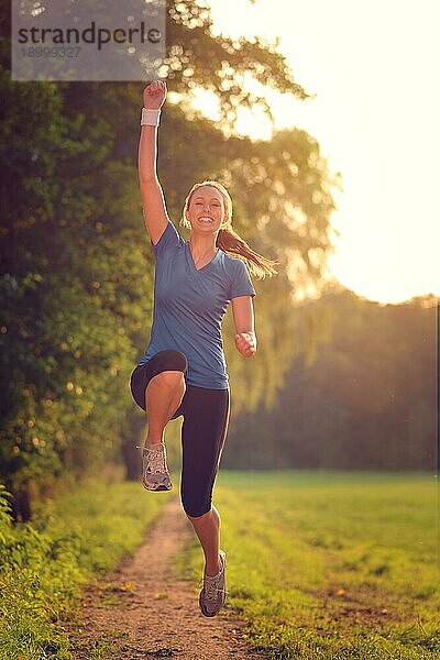 Energetische Frau  die mit einem glücklichen Lächeln voller Vitalität in die Luft springt  während sie während eines körperlichen Trainings auf einem Feldweg läuft