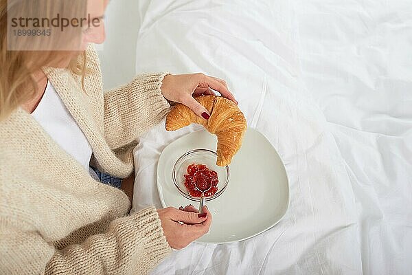 Blick aus der Vogelperspektive auf eine junge Frau  die ein französisches Croissant mit frischer Fruchtmarmelade ißt  während sie ein leckeres Frühstück im Bett genießt