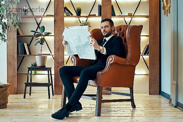 Selbstbewusster junger Geschäftsmann  der morgens im Sessel sitzend in einem modernen Interieur die Zeitung und die neuesten Nachrichten liest
