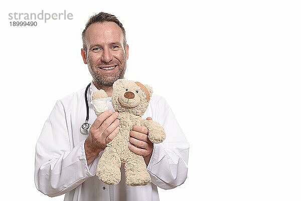 Freundlich lächelnder Kinderarzt mittleren Alters mit unrasierten Bartstoppeln  der einen Teddybär mit bandagiertem Arm hält  um einen jungen Patienten zu beruhigen  vor weißem Hintergrund