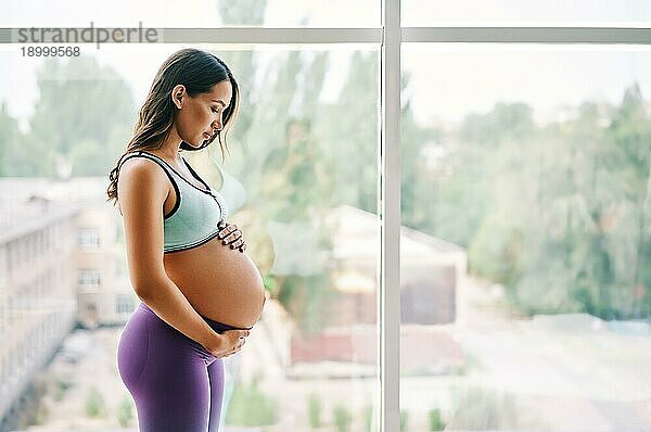 Porträt einer jungen schwangeren Frau in Sportkleidung  die in der Nähe des Fensters steht und ihre Hände auf ihren Bauch legt. Schwangerschaft  Erwartung und Zärtlichkeit Konzept. Bewußte Mutterschaft
