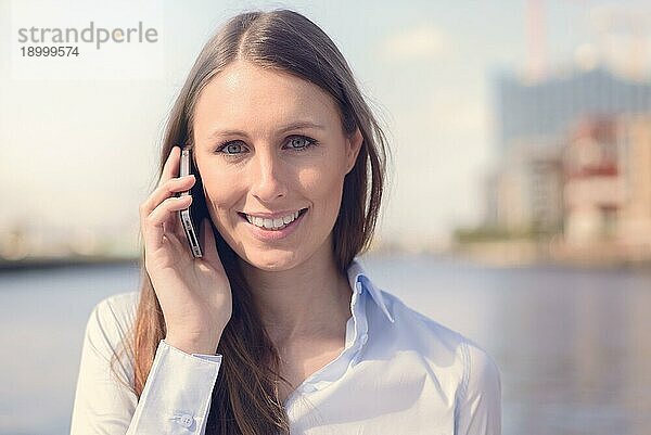 Attraktive Frau  die einen Anruf mit ihrem Mobiltelefon entgegennimmt  steht im Freien vor einem Flusshintergrund und lächelt in die Kamera  während sie dem Gespräch zuhört