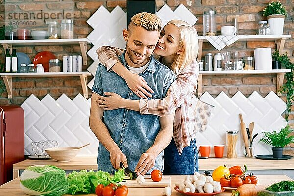 Lächelndes junges Paar kocht zusammen vegetarische Mahlzeit in der Küche zu Hause. Frau umarmt Mann. Gesunder Lebensstil Konzept