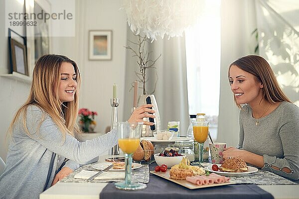 Lächelnde  attraktive junge Teenagerin zeigt ihrer Mutter ihr Handy  während sie gemeinsam zu Hause ein gesundes Frühstück genießen