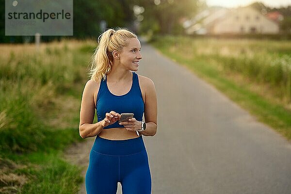 Fitte  sportliche Frau  die beim Joggen auf einem ländlichen Wanderweg ihr Handy konsultiert und dabei mit einem zufriedenen Lächeln zur Seite schaut
