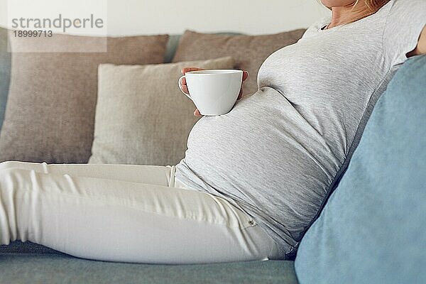 Schwangere Frau entspannt sich auf einem Sofa mit einem großen Becher mit heißem Getränk in einer Nahaufnahme Seitenansicht ihres Babybauchs und Hände halten die Tasse
