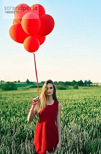 Junge schöne Frau in rotem Kleid posiert im grünen Feld mit roten Luftballons. Freiheit  Spaß  Urlaub Konzept
