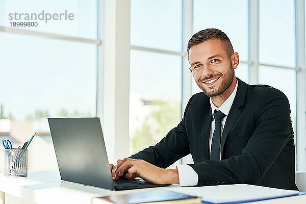 gutaussehender lächelnder Geschäftsmann im eleganten Anzug  der in einem hellen modernen Büro am Laptop arbeitet. geschäftliches Erfolgskonzept