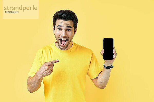Aufgeregter schreiender Mann  der mit dem Finger auf sein Handy zeigt und in die Kamera schaut  auf gelbem Studiohintergrund