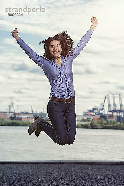 Junge Frau  die mit erhobenen Armen und einem triumphierenden Lächeln vor dem Hintergrund einer Wasserstraße und einer entfernten Stadt vor Freude springt und jubelt