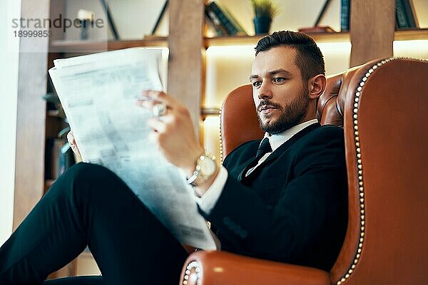 Selbstbewusster junger Geschäftsmann  der morgens im Sessel sitzend in einem modernen Interieur die Zeitung und die neuesten Nachrichten liest