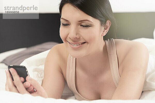 Attraktive junge Frau im Bett liegend  die eine SMS auf ihrem Handy liest und dabei genüsslich über die Nachricht lächelt