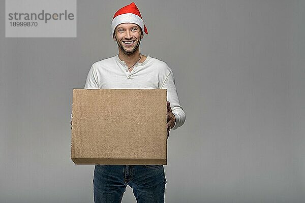 Fröhlicher attraktiver junger Mann mit Weihnachtsmannmütze  der einen großen braunen Karton für Weihnachten trägt oder ausliefert  über grauem Hintergrund mit Leerzeichen