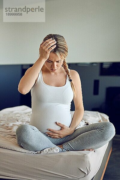 Besorgte hochschwangere junge Frau  die im Schneidersitz auf einem Bett sitzt  ihren geschwollenen Bauch stützt und sich an den Kopf faßt und mit ernster Miene nach unten schaut