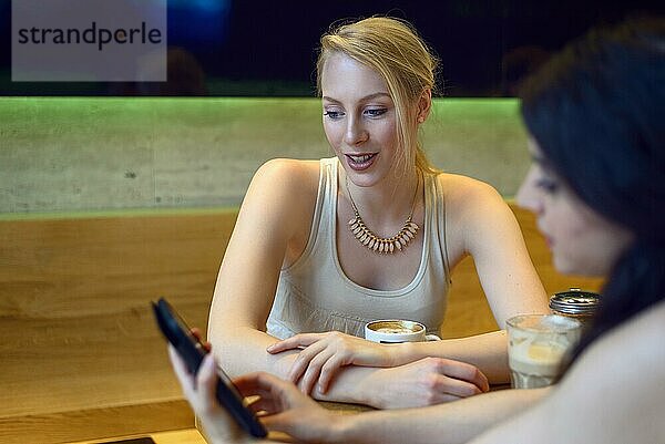 Zwei junge Frauen schauen auf ein digitales Tablet  während sie in einem Bistro oder einer Kaffeebar zusammensitzen und einen Kaffee genießen