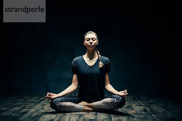 Junge attraktive Frau übt Yoga sitzen in Lotuspose und meditieren in dunklen Raum. Ardha Padmasana Übung. Gleichgewicht und Erholung Konzept