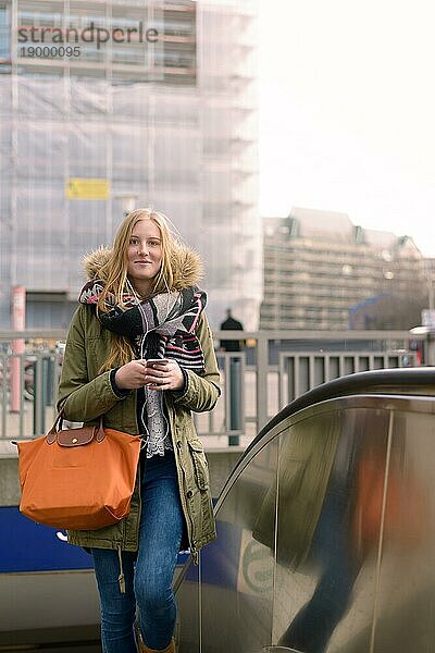 Stilvolle blonde Frau zu Fuß in Winter Stil Mode  während er sein Handy mit ihrem eleganten Orange Ledertasche auf ihrem Arm