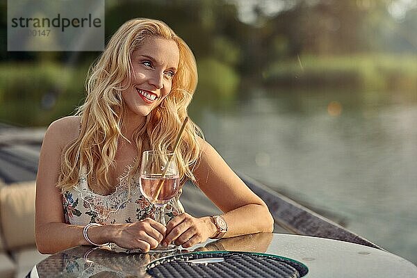 Glückliche junge Frau  die sich mit einem Cocktail zum Sundowner an einem Tisch im Freien auf einer Holzterrasse mit Blick auf ruhiges Wasser entspannt und mit einem Lächeln des Vergnügens zur Seite schaut und einen warmen Sommerabend genießt