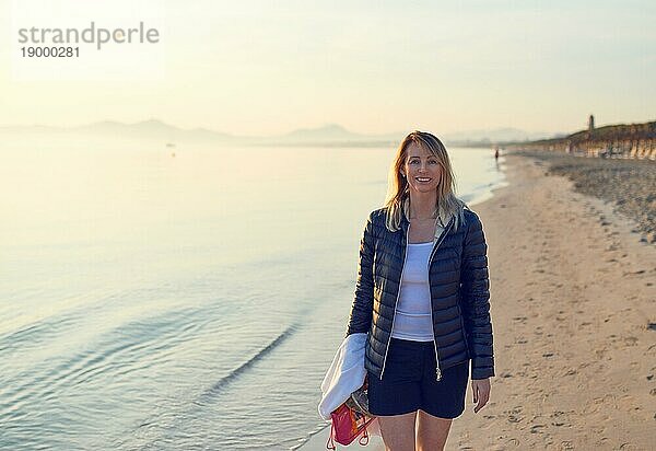 Fröhliche blonde Frau geht bei Sonnenuntergang am Strand entlang