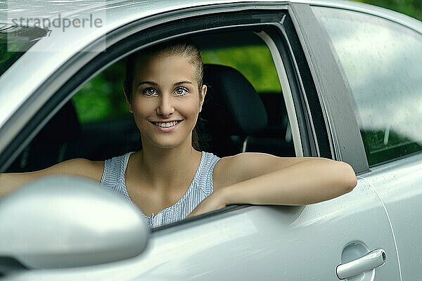 Lächelnde attraktive junge Frau am Steuer eines Autos  die mit einem warmen freundlichen Lächeln aus dem offenen Seitenfenster in die Kamera schaut