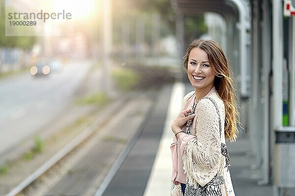 Attraktive  stilvolle junge Frau  die allein auf dem Bahnsteig eines kleinen städtischen Bahnhofs wartet und mit einem glücklichen Lächeln in die Kamera blickt  Kopiervorlage nach links