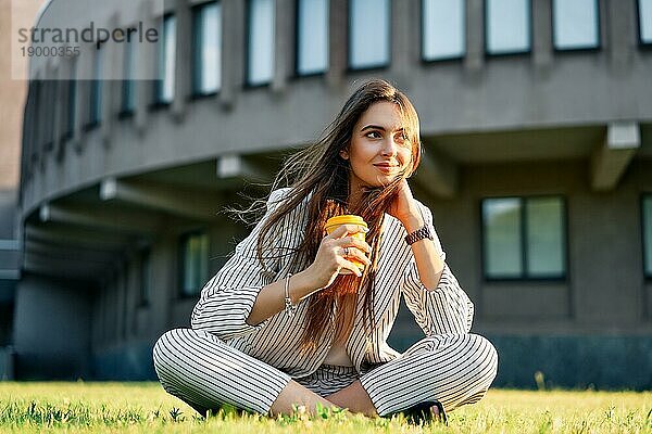 Junge stilvolle Frau trinkt Kaffee und entspannen Sie sich auf einem Rasen im Stadtpark sitzen. Pause Zeit Konzept