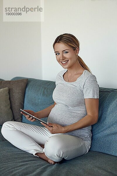 Glückliche schwangere Frau  die sich auf einem Sofa entspannt  im Schneidersitz ein eBook auf einem Tablet liest und mit einem warmen  freundlichen Lächeln in die Kamera schaut